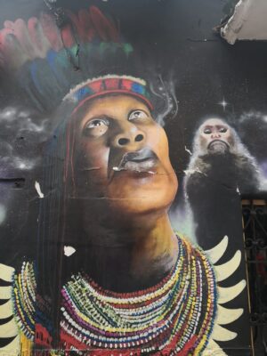 This is a colorful mural depicting an indigenous man with a headdress and necklace, smoking, with a monkey in the background, against a starry night sky.